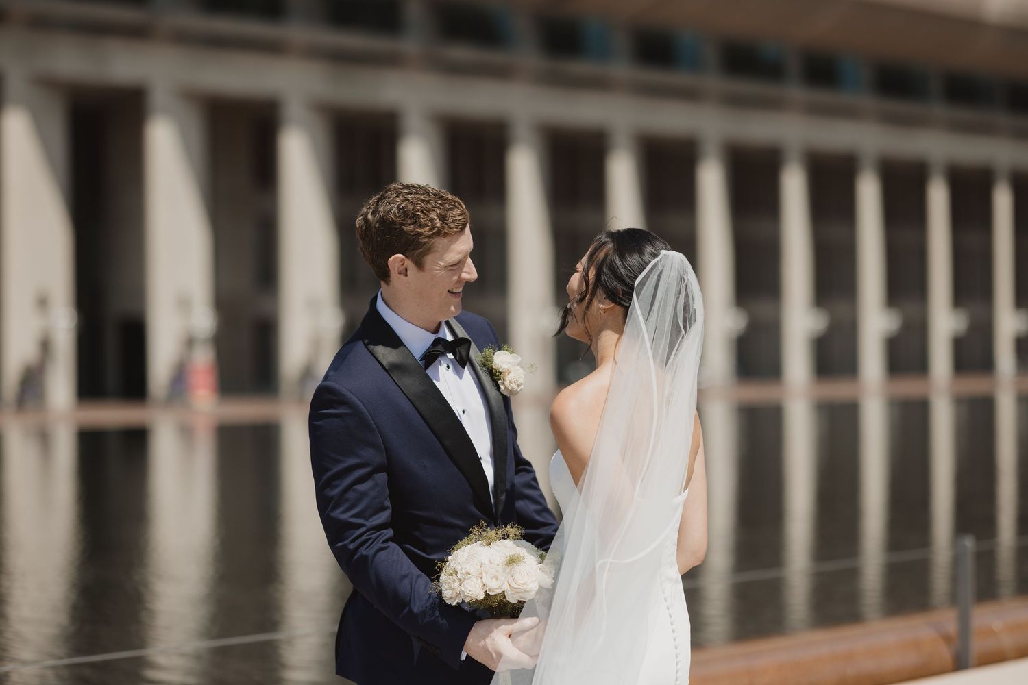 Bride and groom share a romantic moment outdoors, she holds white bouquet, he wears navy tuxedo with bow tie.