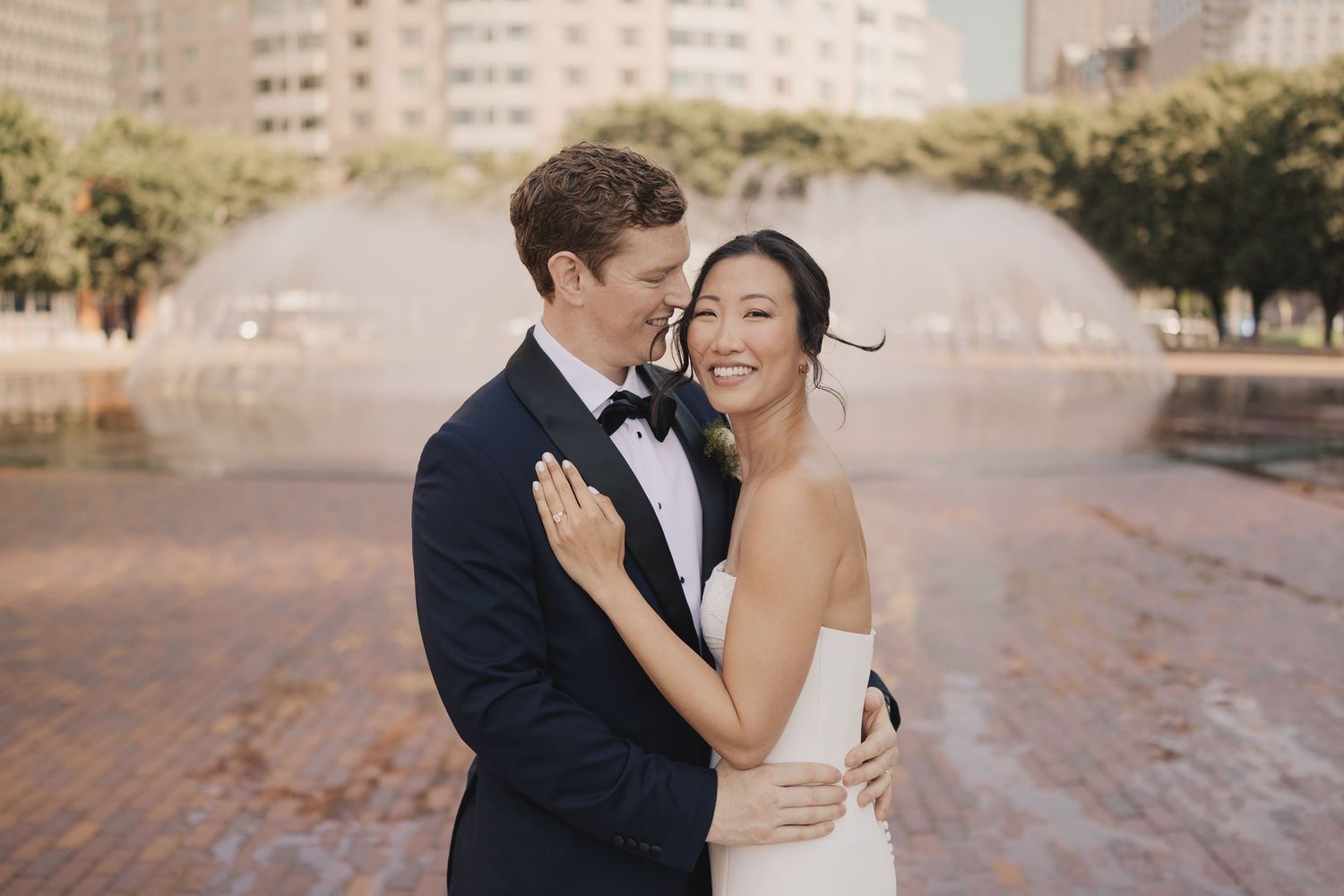 Bride and groom embrace smiling in front of a fountain in an urban setting, groom in tuxedo, bride in strapless white gown.