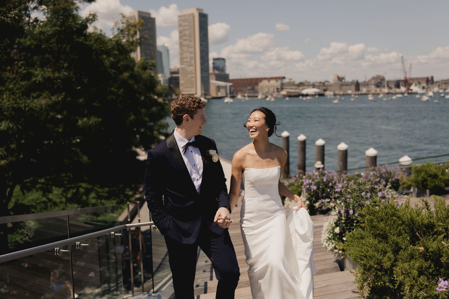 Smiling bride and groom hold hands walking along waterfront boardwalk with city skyline on sunny wedding day.