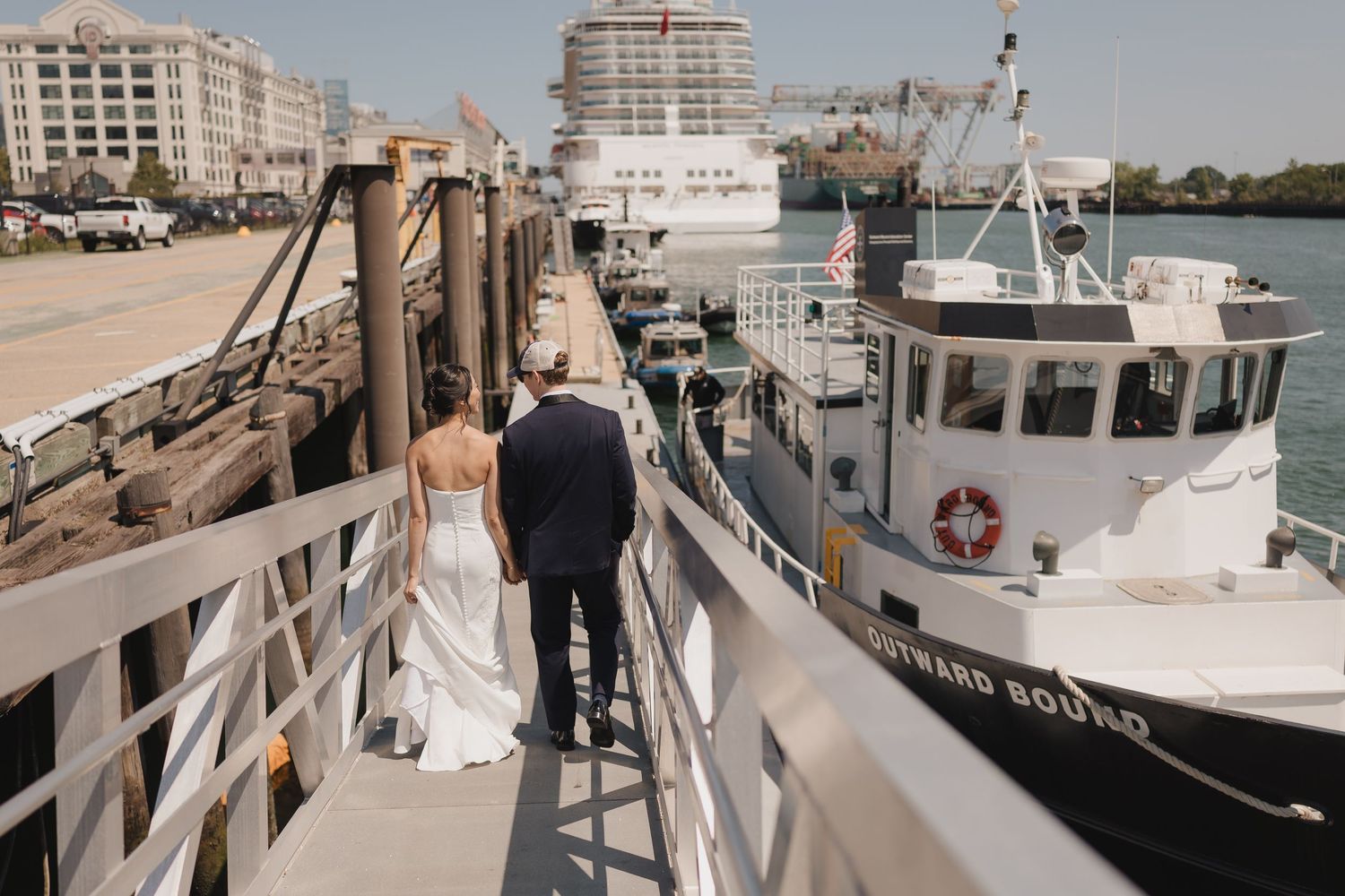 Bride and groom walk hand in hand down a dock toward a boat on their wedding day, bride in flowing white gown.
