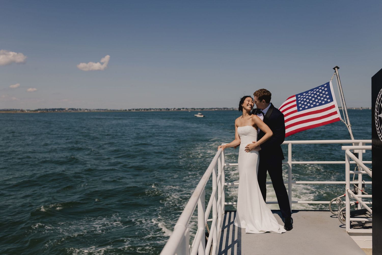 Bride and groom share a romantic moment on a yacht deck with an American flag and open water behind them.