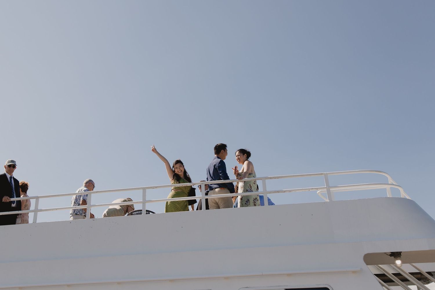 Wedding guests enjoy views from the upper deck of a yacht on a sunny clear-sky day.