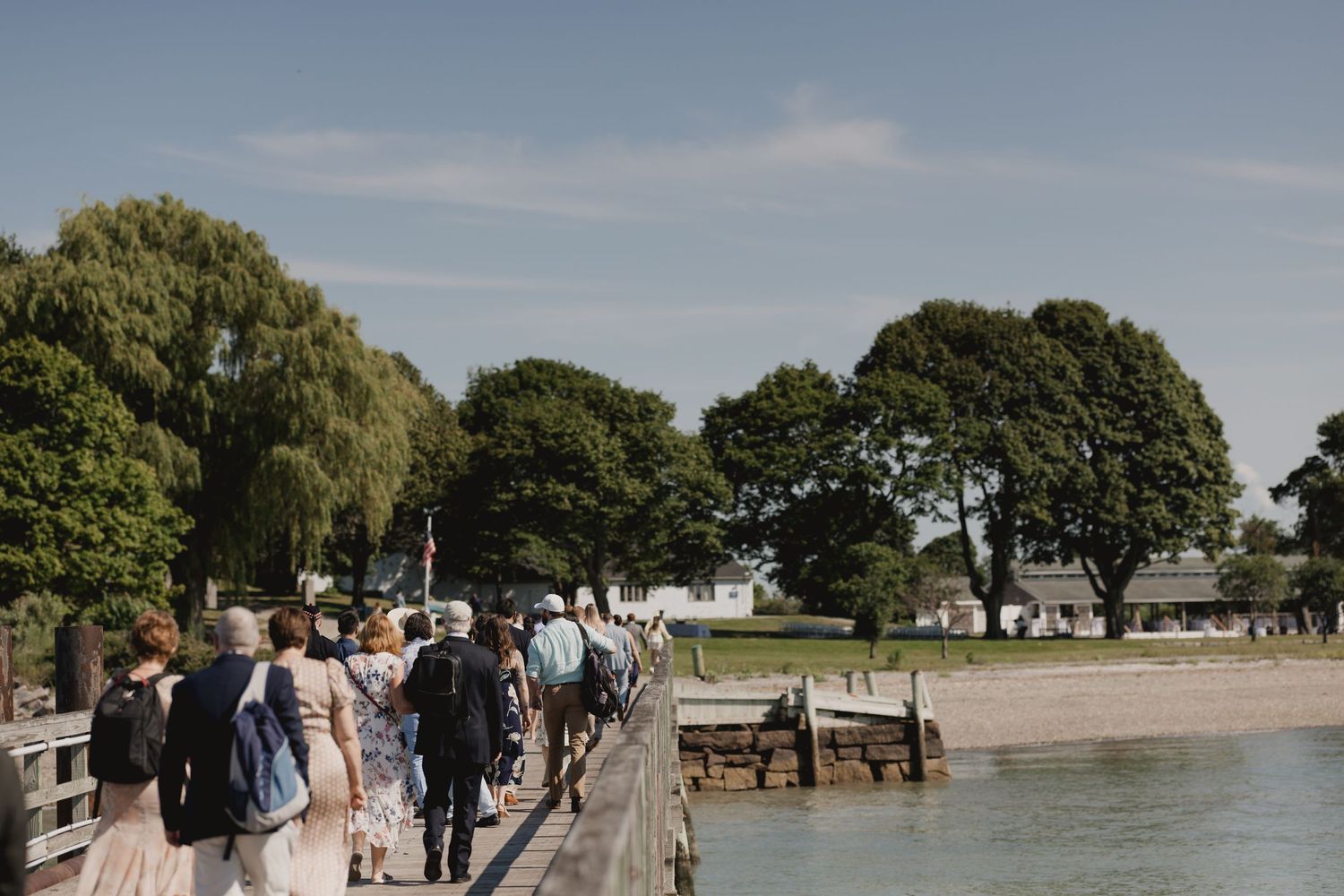 Wedding guests walk along a wooden pier beside calm water surrounded by lush green trees on a sunny day.