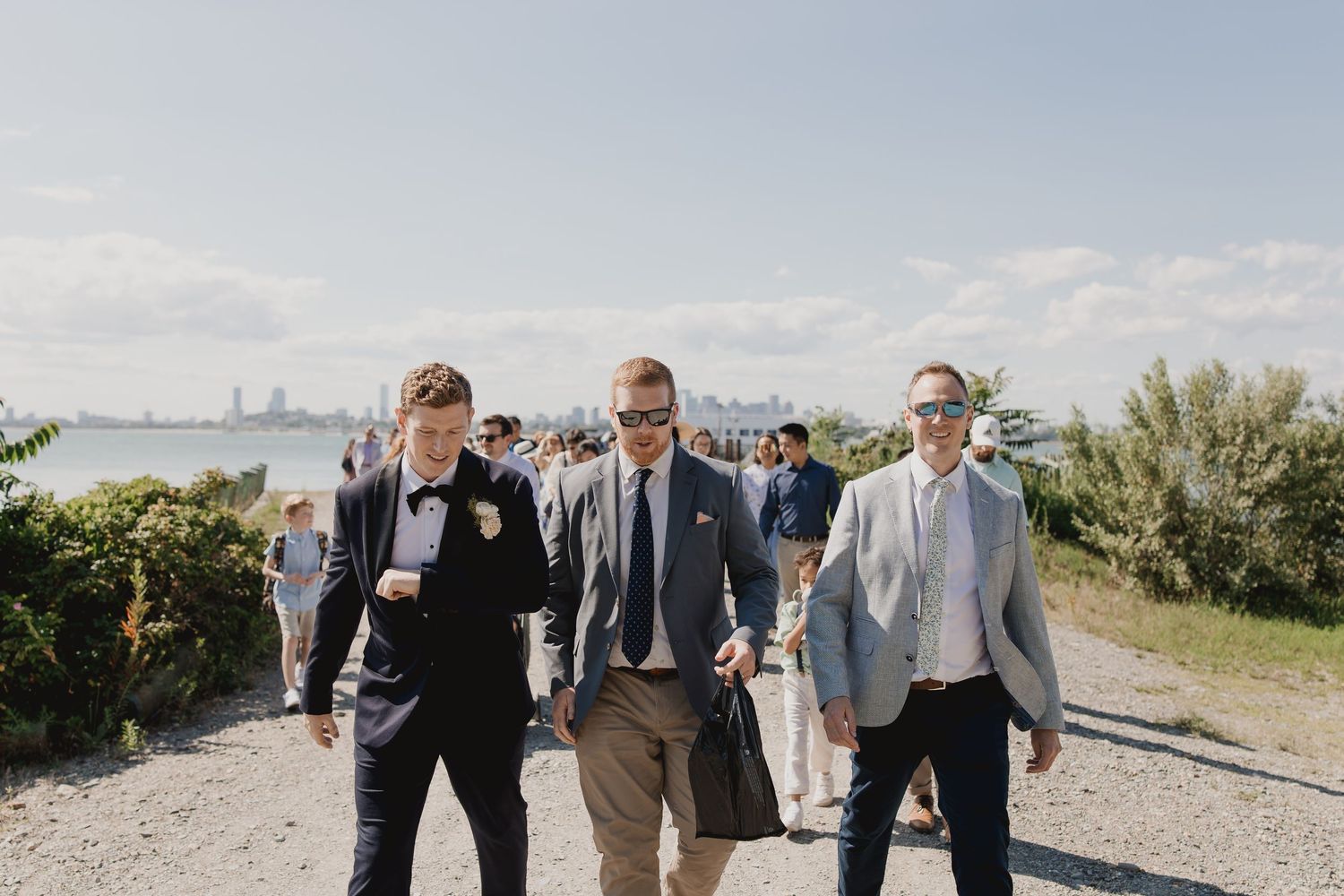Three well-dressed men in suits walk together outdoors at a waterfront wedding celebration.