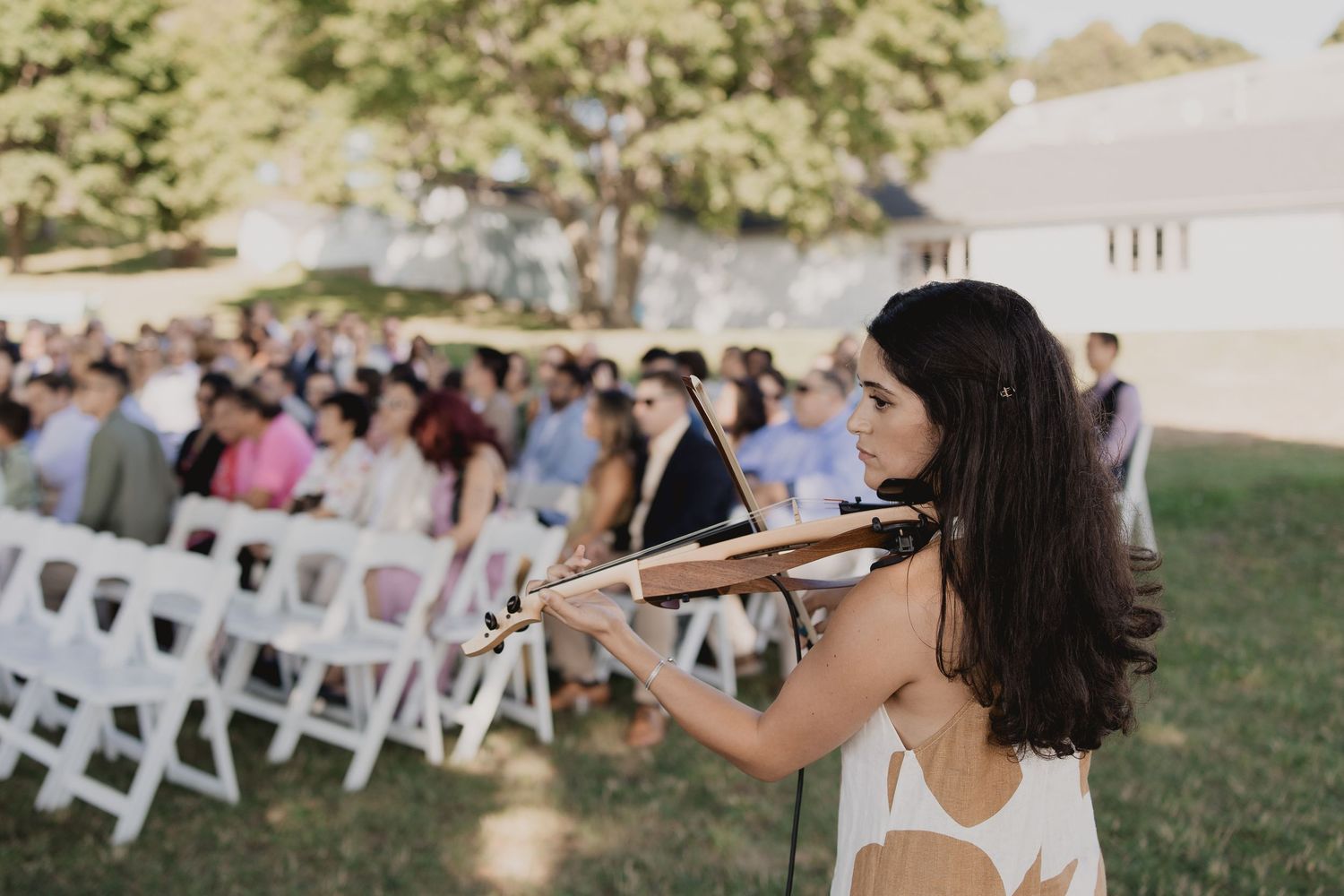 A violinist performs live music for wedding guests seated in white chairs during an outdoor ceremony on a sunny day.