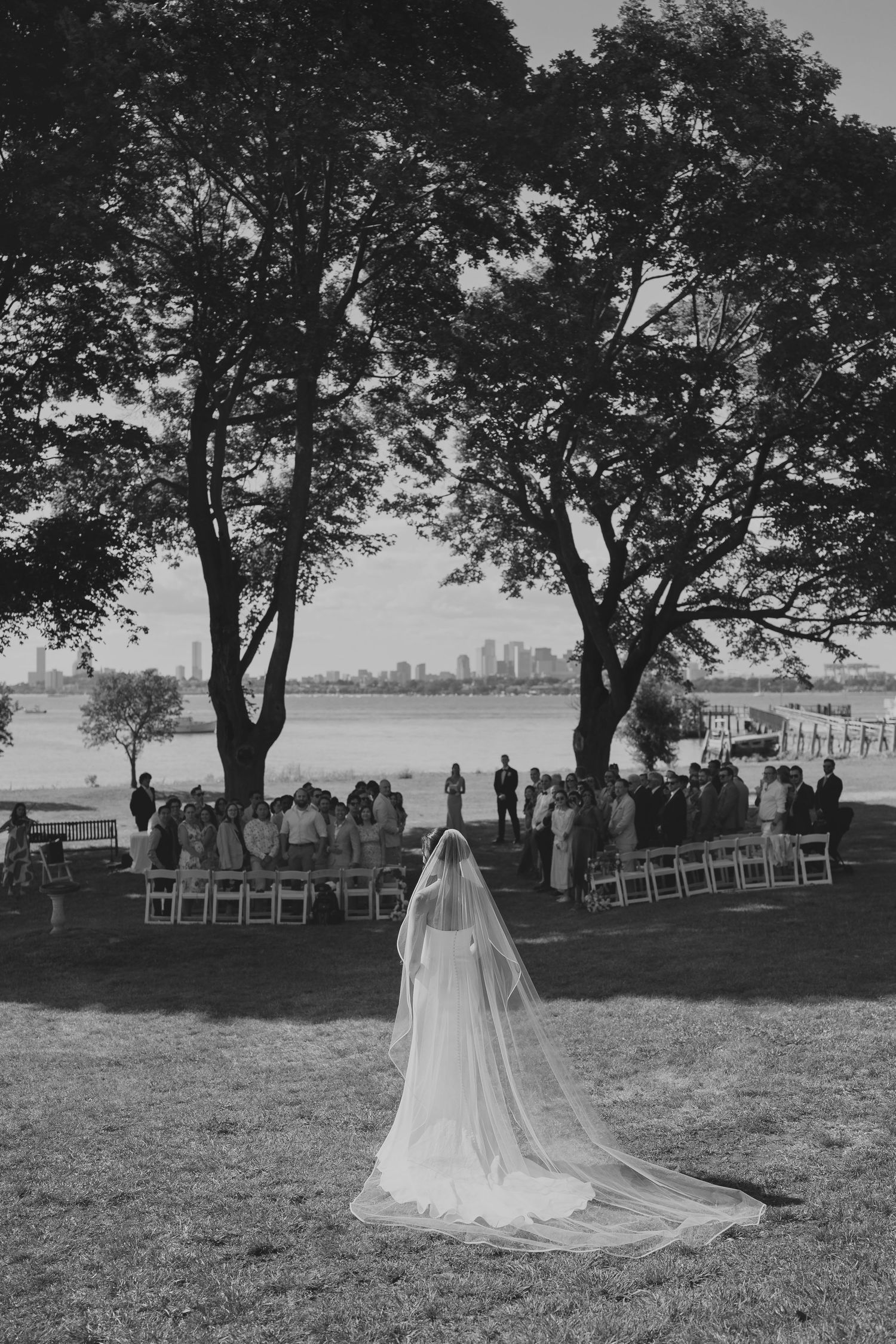 Black and white photo of a bride in a veil walking toward an outdoor waterfront wedding ceremony.