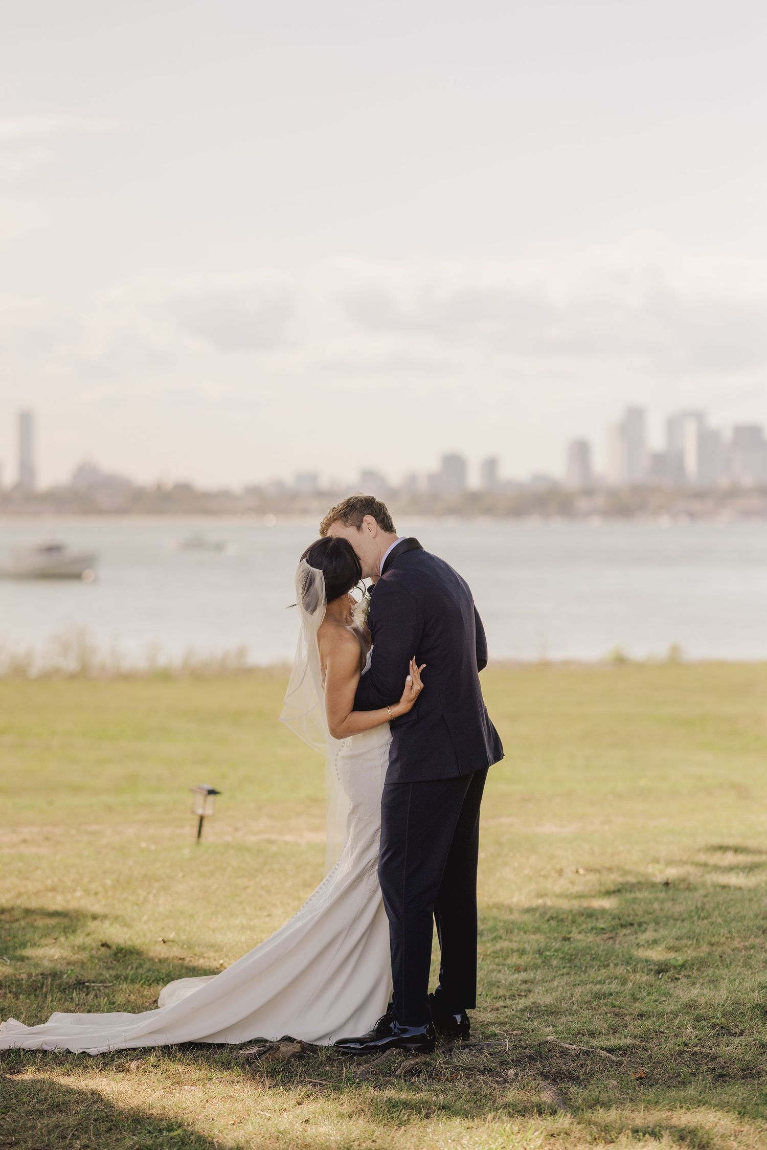 Bride and groom share a tender embrace on a lawn with a city skyline and water in the background.
