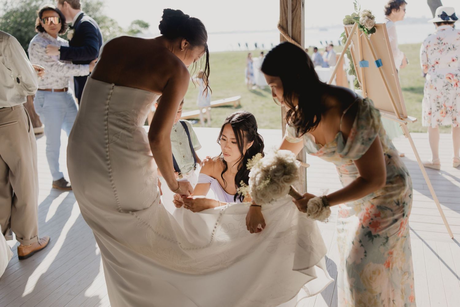 Bridesmaids help a smiling bride adjust her white wedding gown at an outdoor venue.