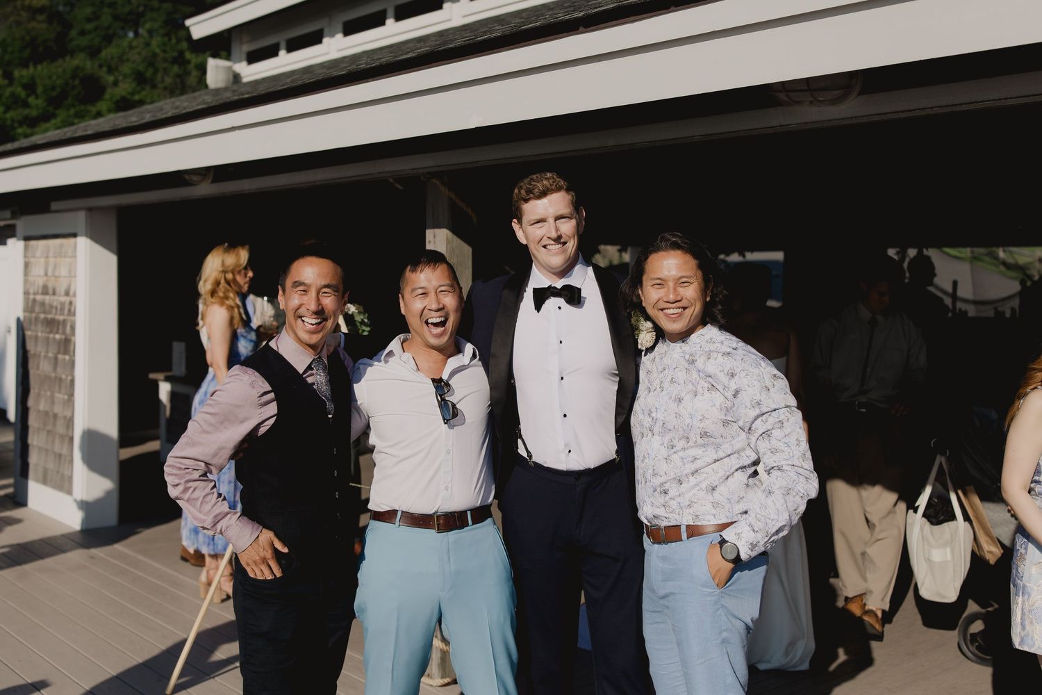 Groom in black tuxedo smiles with three friends outdoors at wedding celebration.