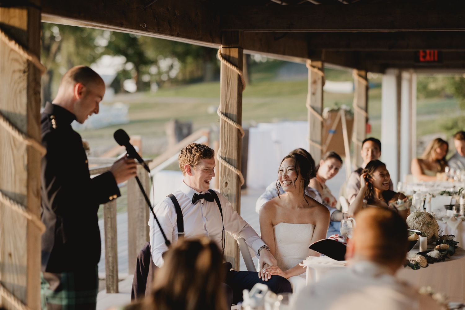 A military groom gives a speech as the bride smiles during a wedding reception under string lights.