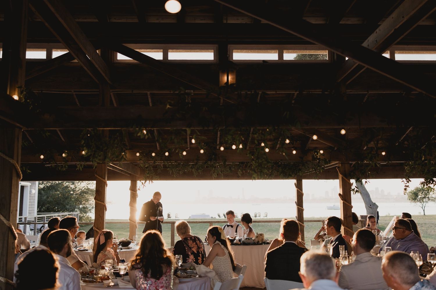 Wedding guests dine under string lights in a rustic open-air venue with waterfront views at sunset.