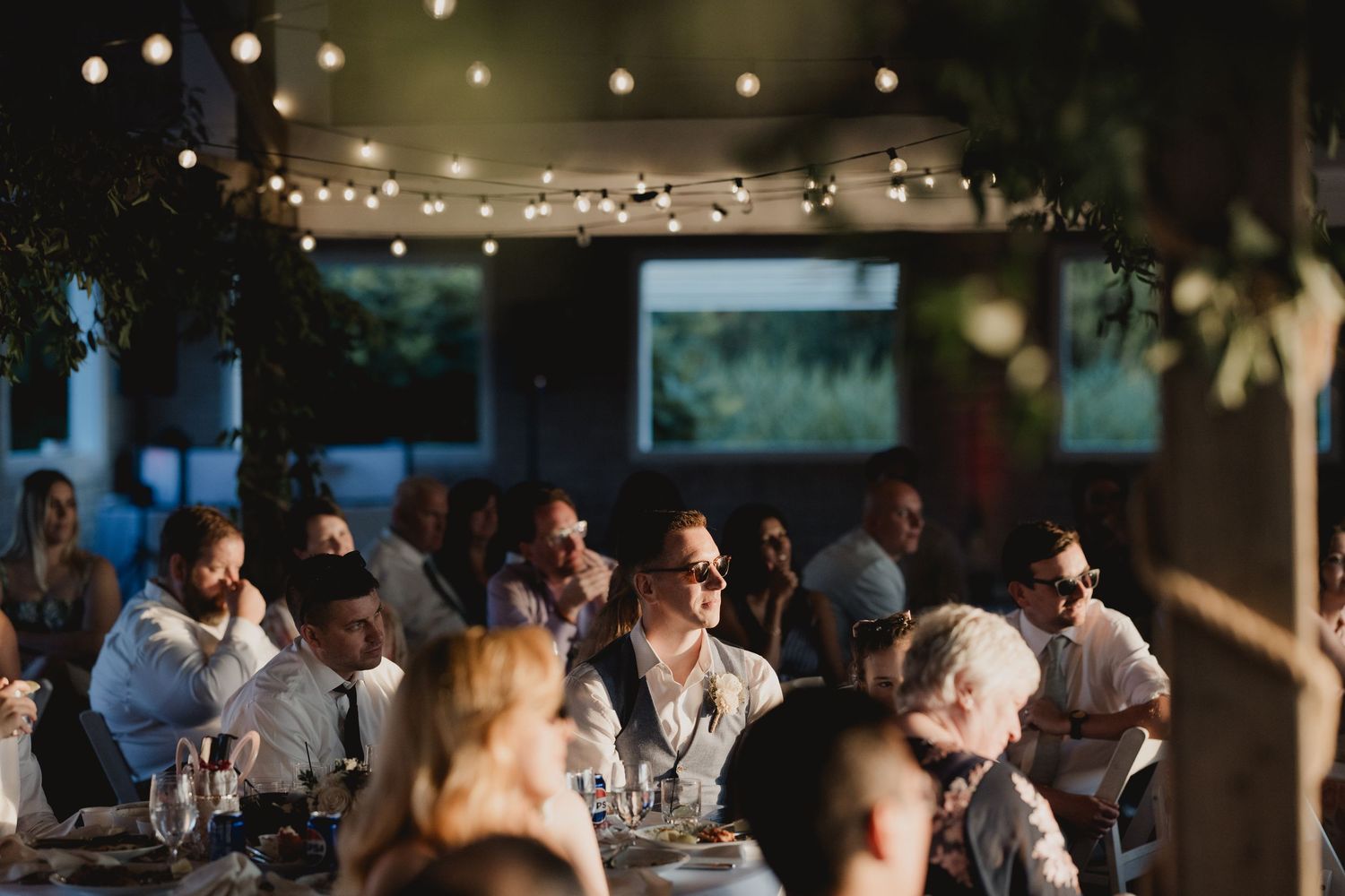 Wedding guests seated at decorated tables enjoy the reception under warm string lights in a dimly lit venue.