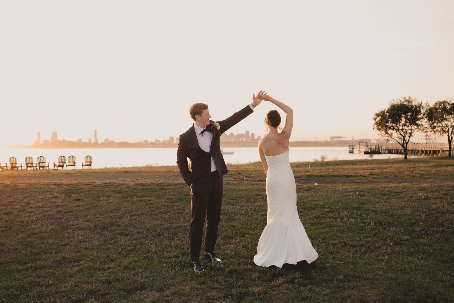 Groom twirls bride in white gown at sunset with city skyline backdrop during outdoor wedding celebration.