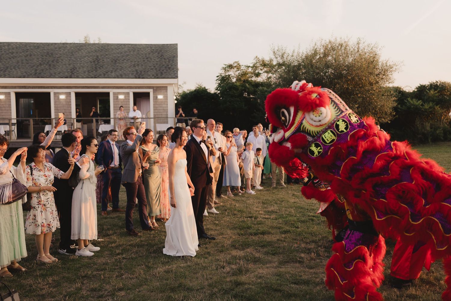 Wedding guests cheer as a vibrant red Chinese lion dancer performs for the newlyweds outdoors.
