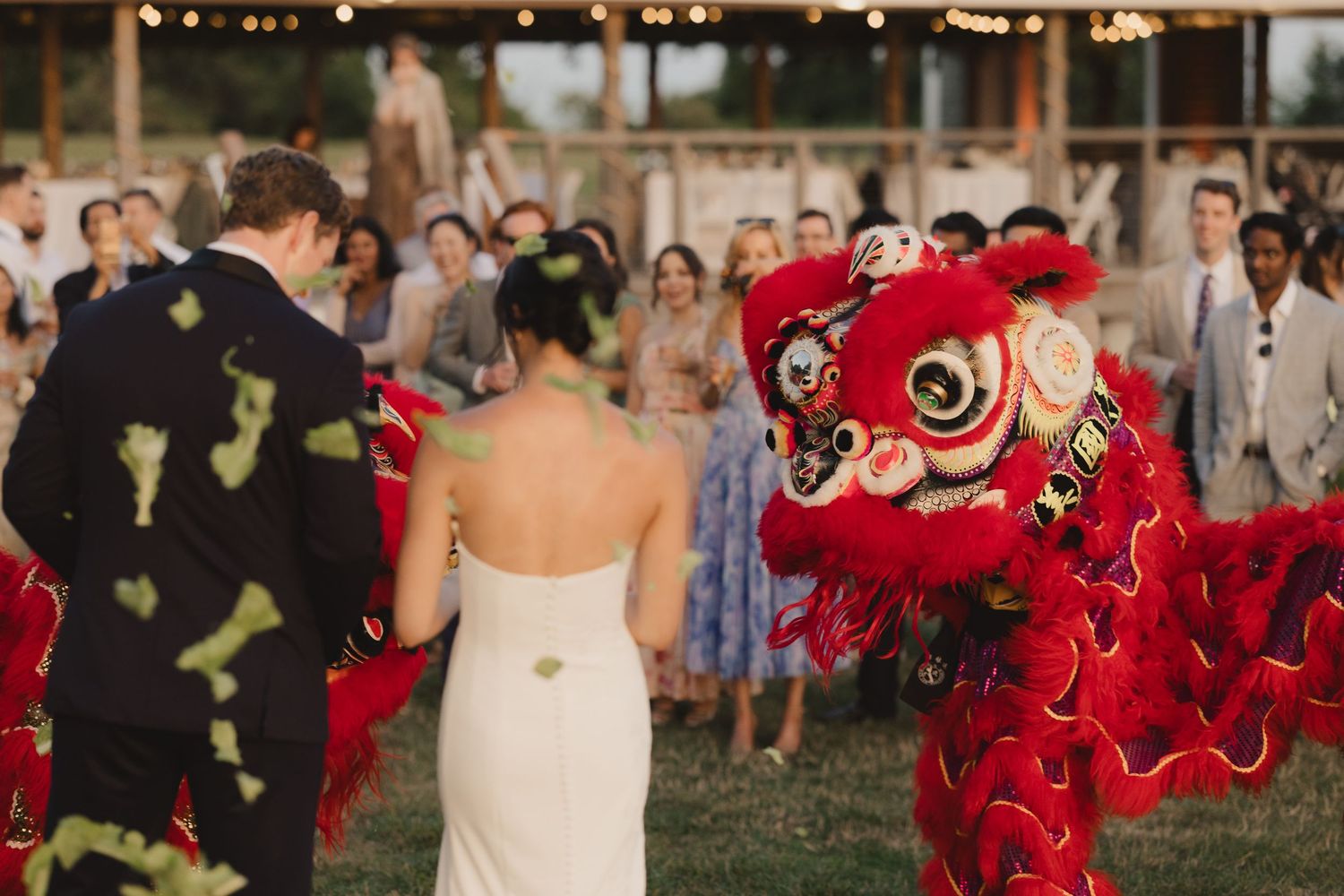 A vibrant red Chinese lion dance performer entertains the bride and groom at an outdoor wedding reception.