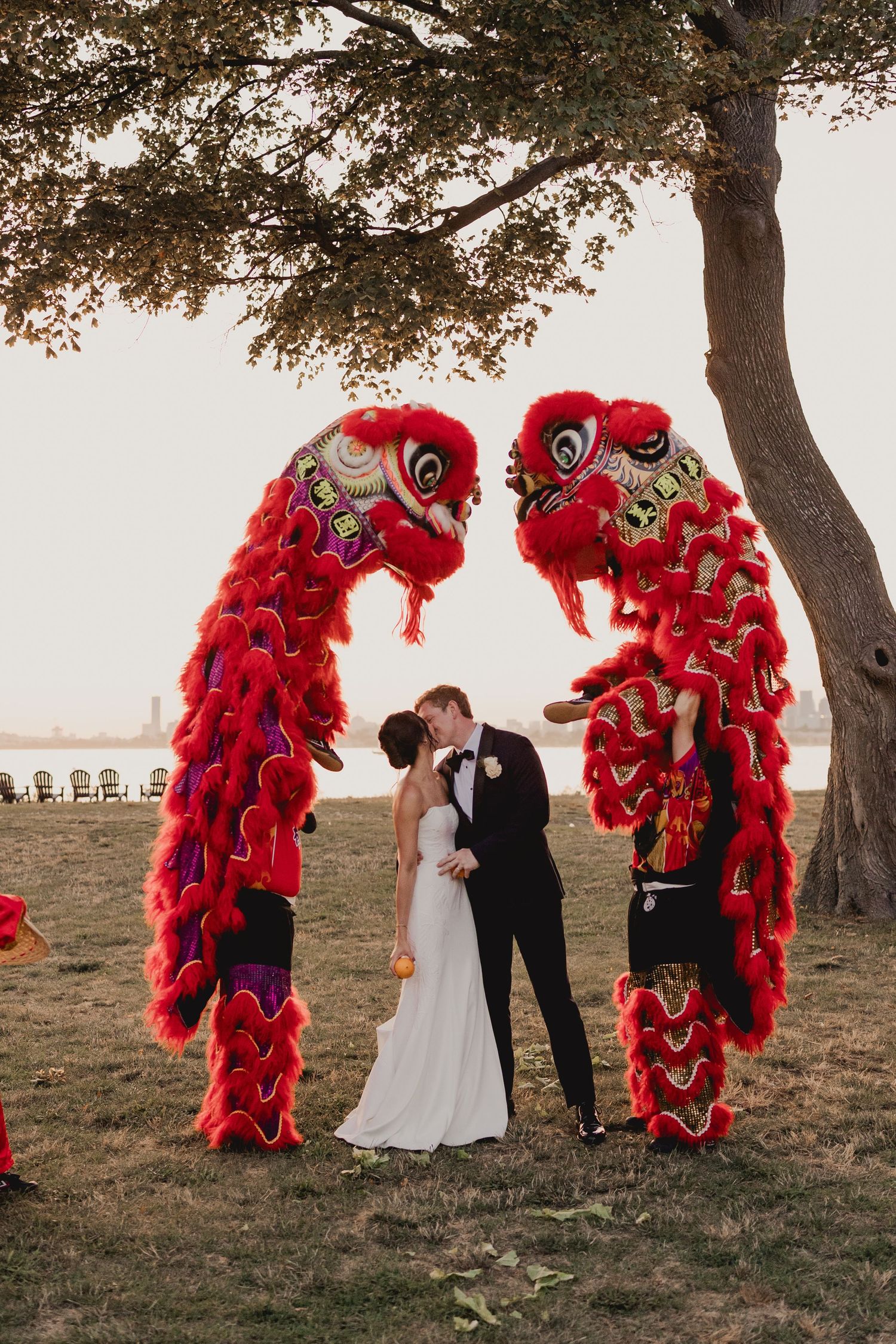 Bride and groom share kiss framed by two vibrant red Chinese lion dance costumes during outdoor wedding.