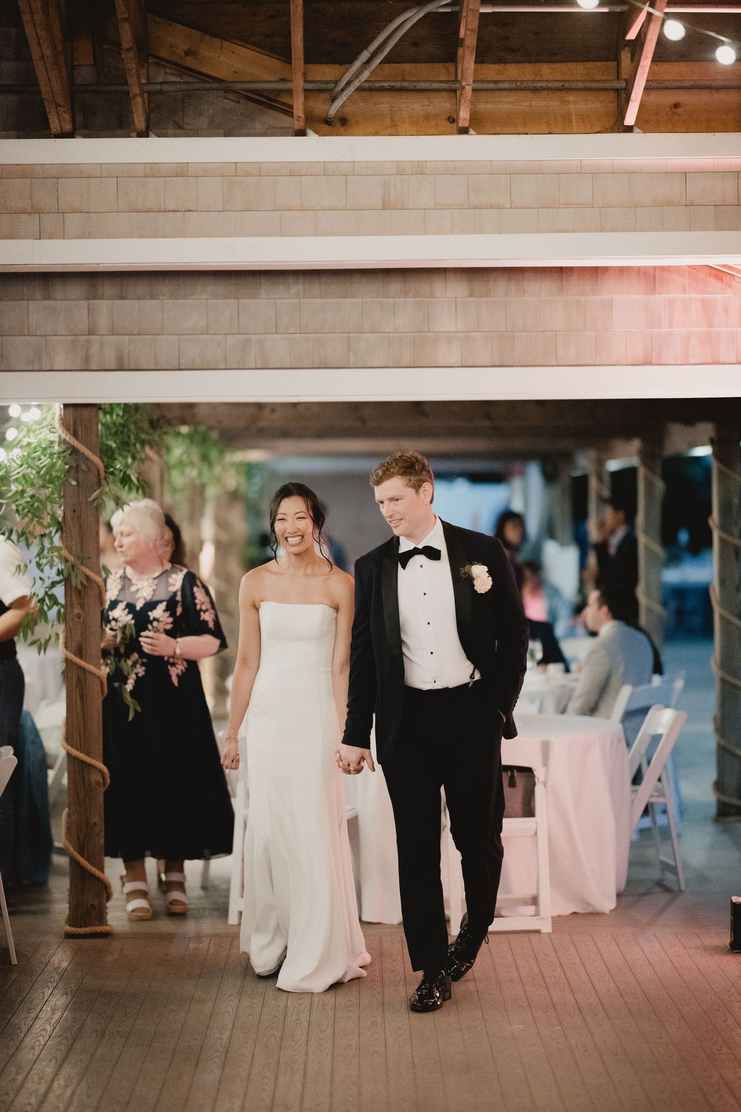 Bride in white gown and groom in black tuxedo hold hands making grand entrance at elegant wedding reception.