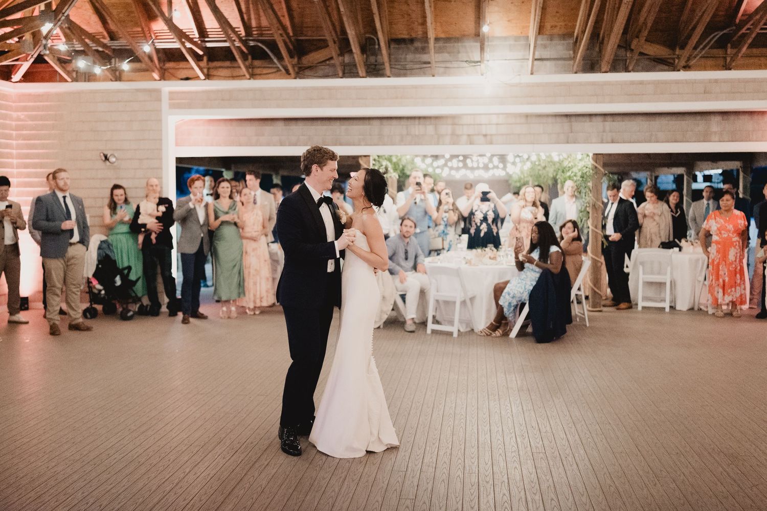Bride and groom share first dance in rustic wood-beamed reception hall surrounded by seated wedding guests.