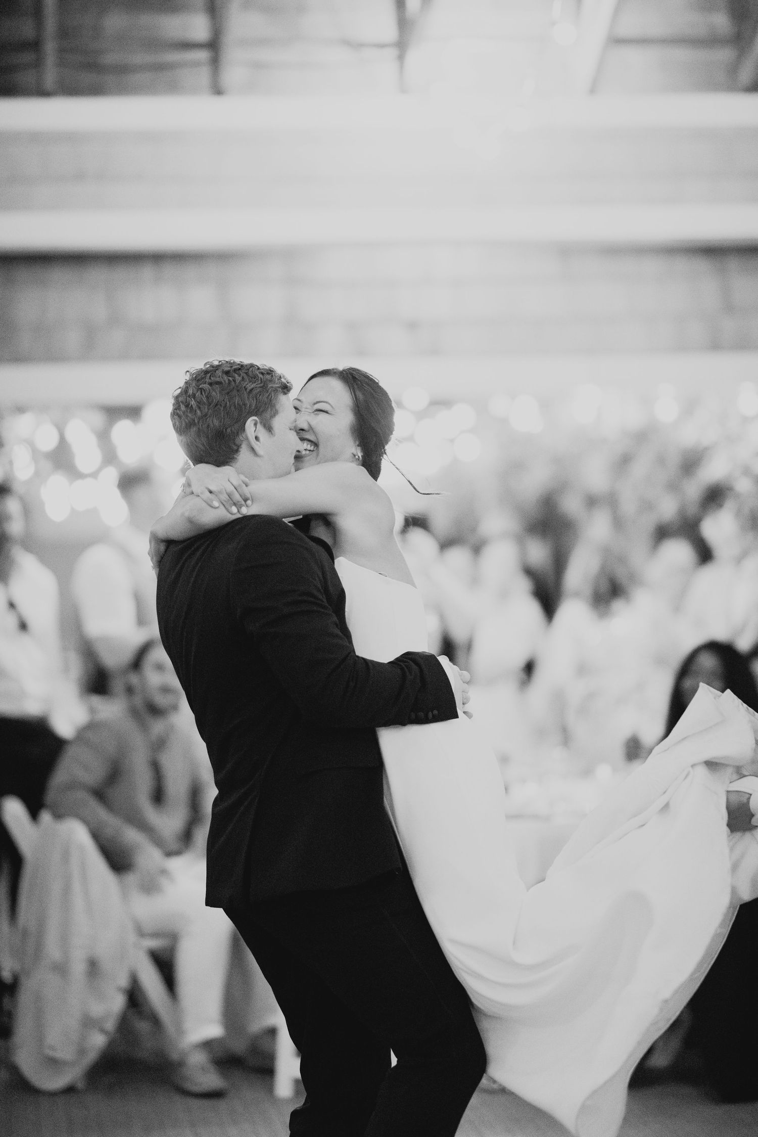 Black and white photo of a laughing bride being lifted by her groom during their first dance at the reception.