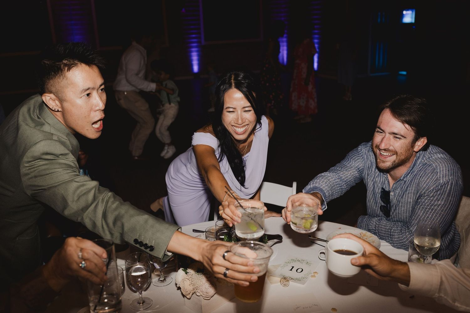 Three friends laugh and clink glasses together at a wedding reception table under purple ambient lighting.