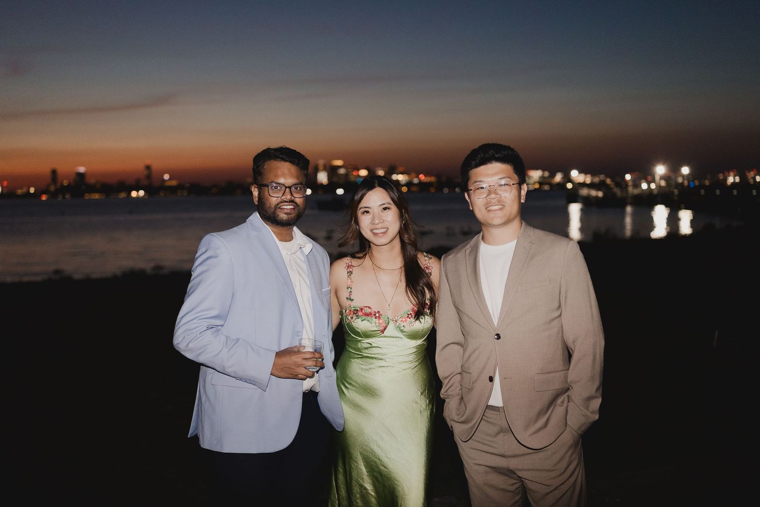 Three wedding guests pose together at twilight with glowing city skyline reflected on water behind them.