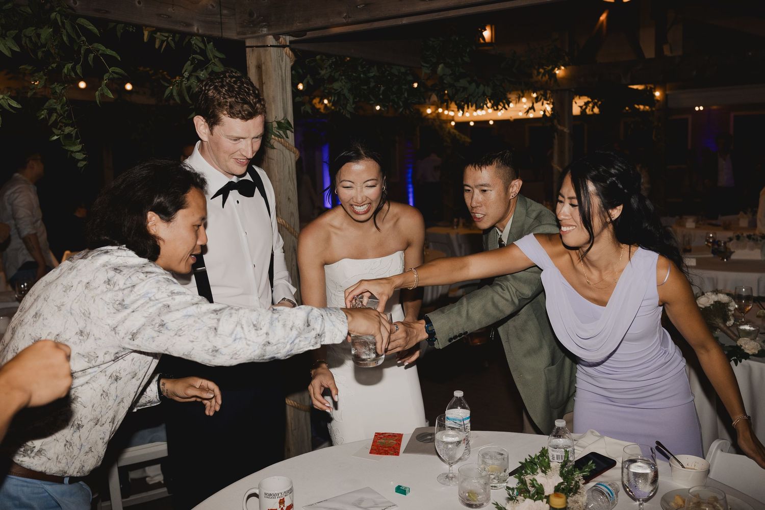 Wedding guests gather around couple at reception table for a celebratory toast under string lights.