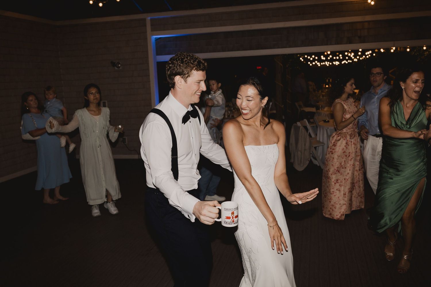 Bride in white gown and groom in suspenders dance joyfully with guests at lively wedding reception.