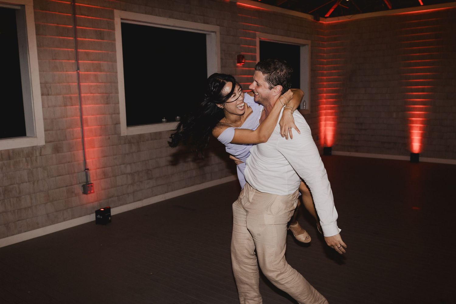 Groom joyfully lifts bride on the dance floor with warm amber uplighting at wedding reception.