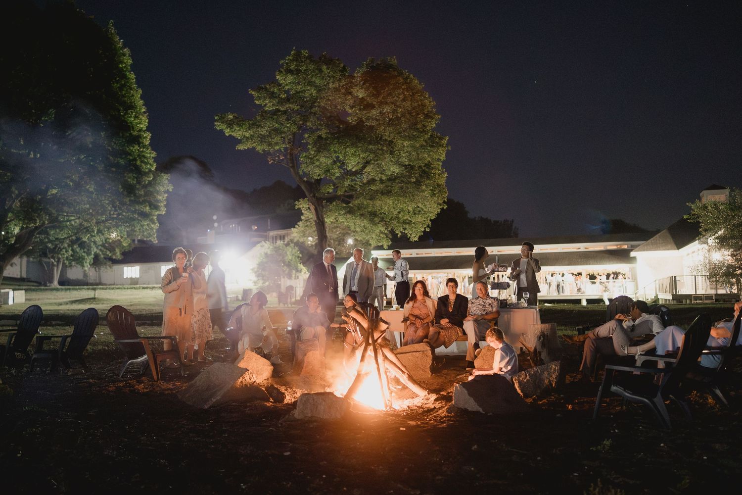Wedding guests gather around a glowing bonfire under the night sky at an outdoor reception.