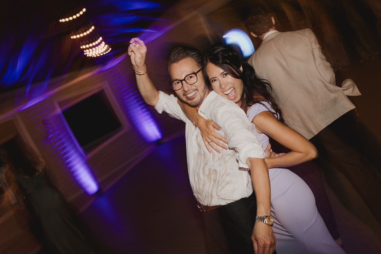 Smiling woman in white dress dances with man in glasses under purple lights at wedding reception.