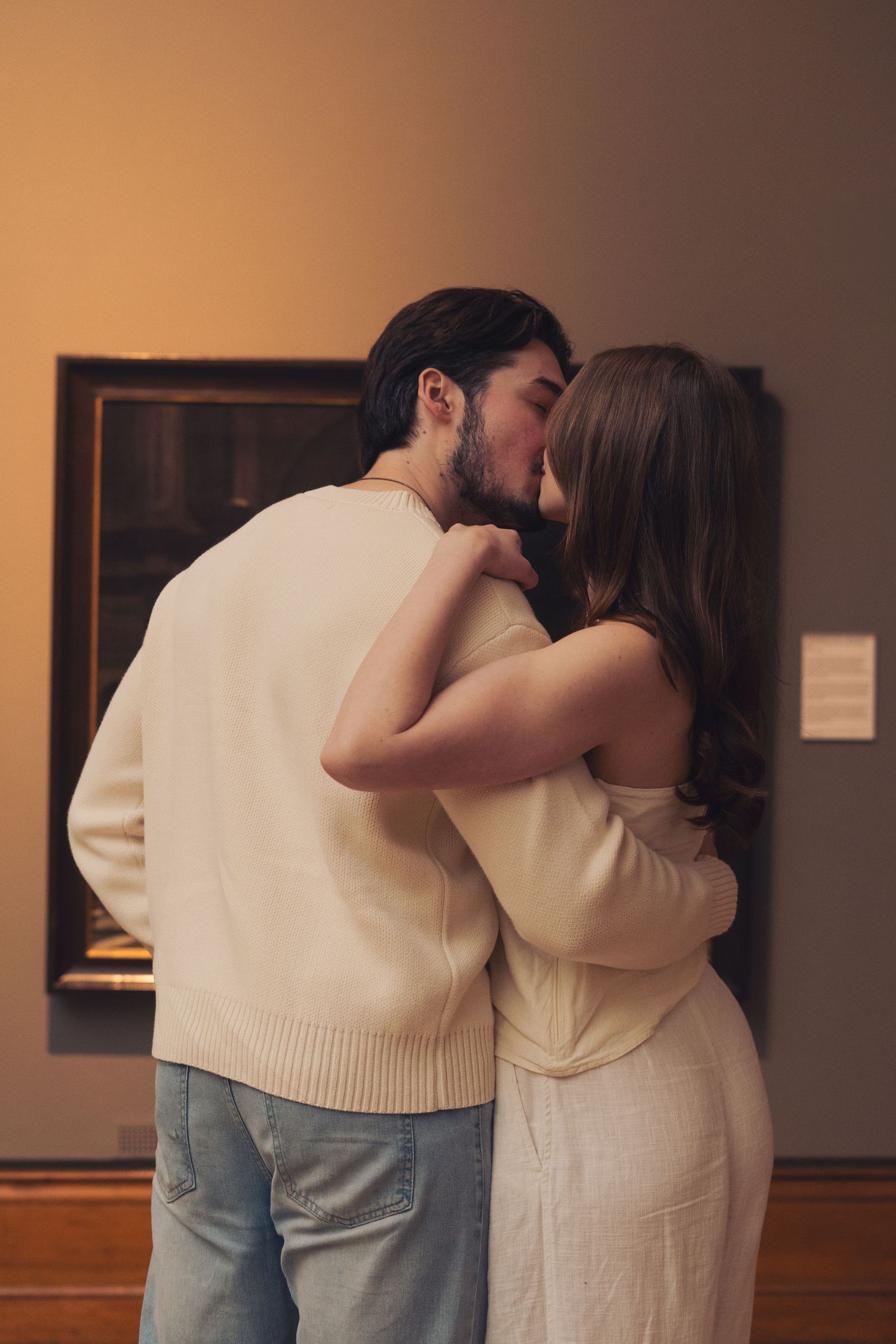 Couple holding hands while viewing classic paintings together in a warmly lit art museum gallery.
