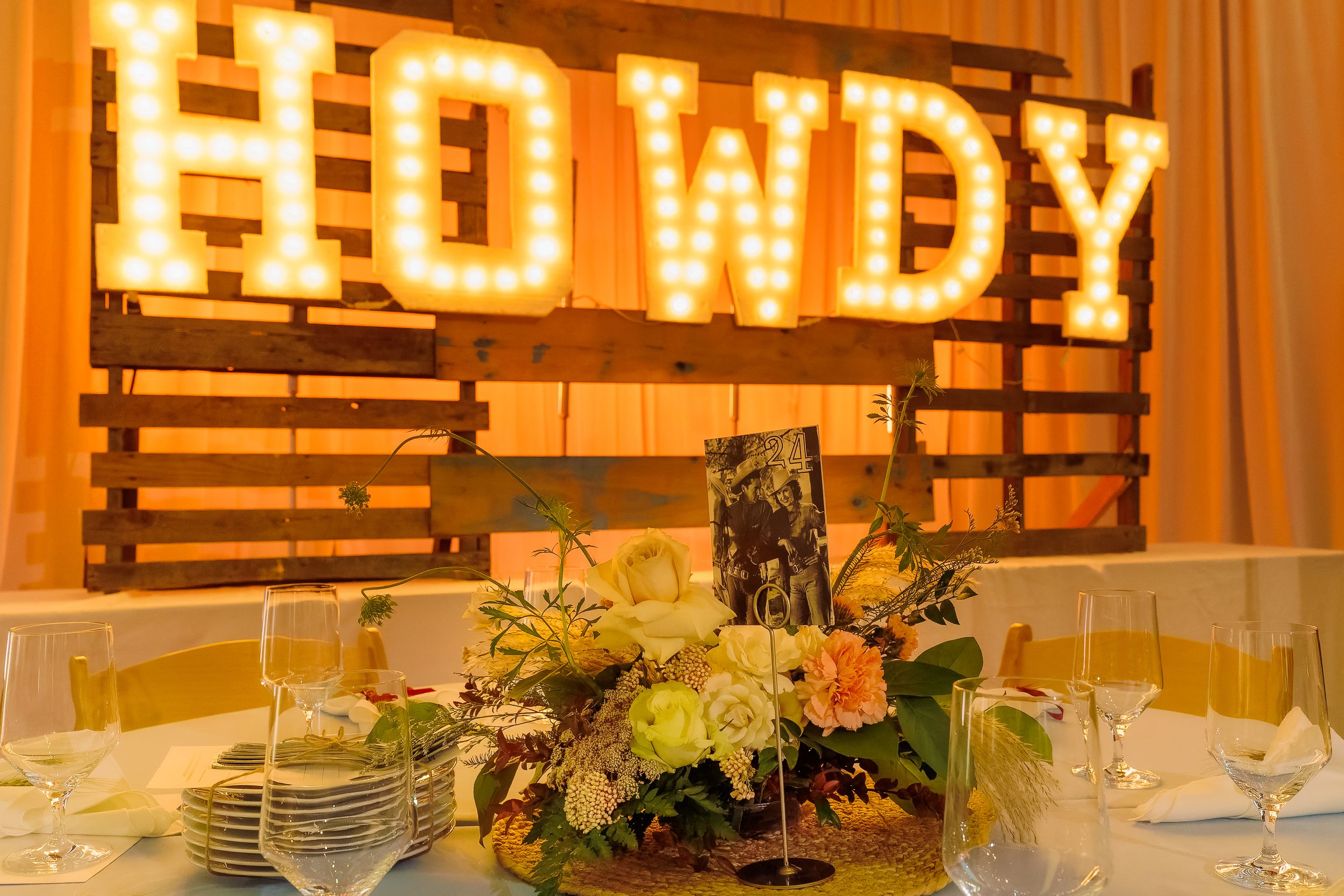 Rustic wedding reception with illuminated HOWDY marquee sign on pallet backdrop and floral centerpiece on decorated table.
