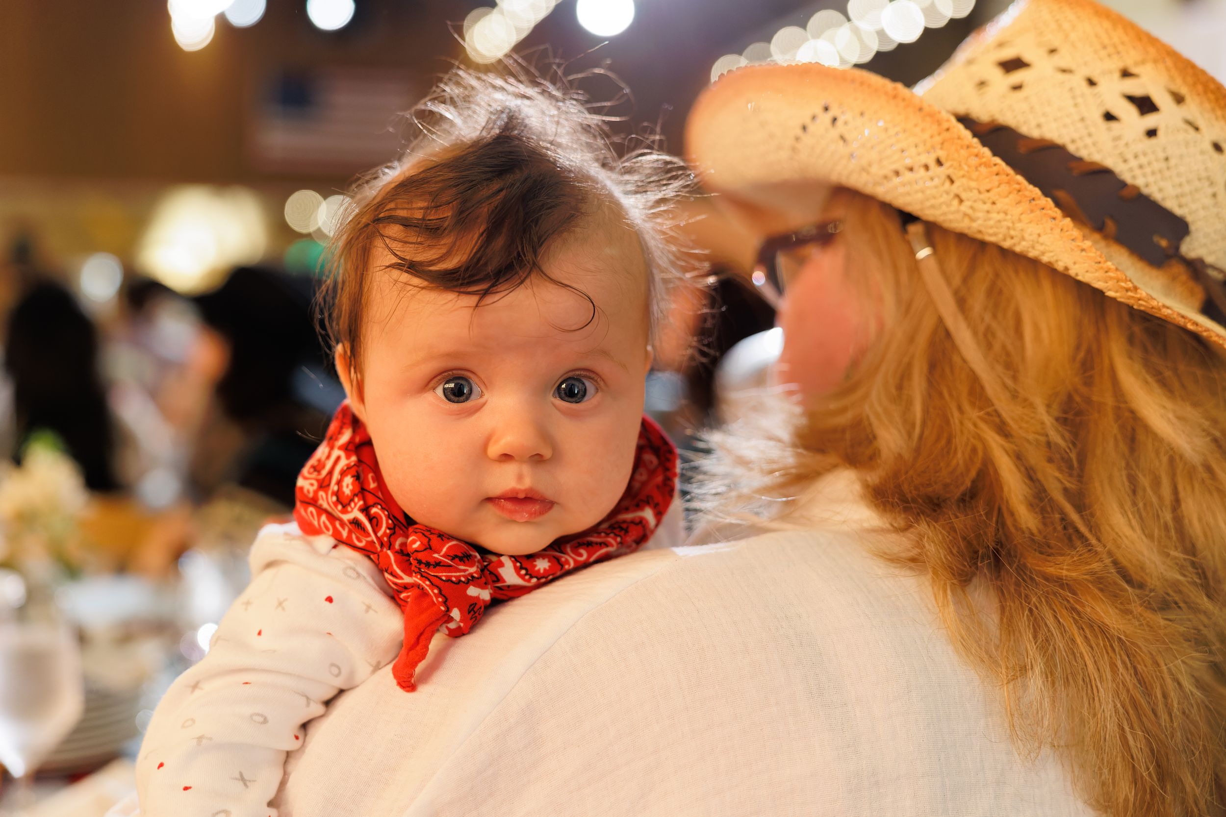 A woman in a straw cowboy hat holds a baby wearing a red bandana at an indoor event.