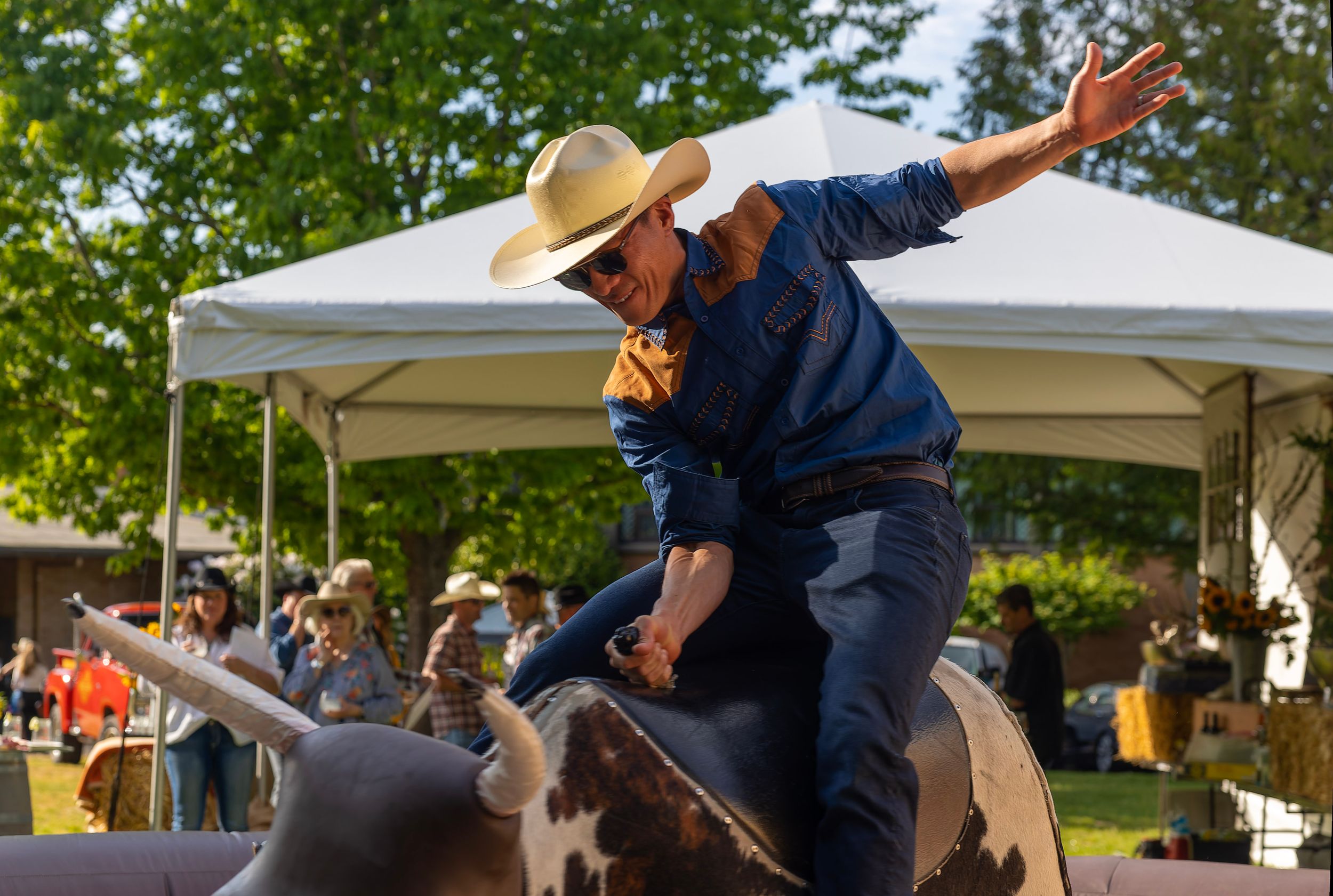 Man in cowboy hat riding a mechanical bull at an outdoor rodeo event under a white tent.