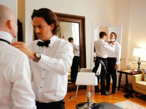 Formal wedding preparations in a bright room with tuxedos and bow ties being adjusted.