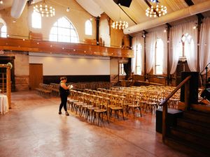 Large banquet hall with wooden chairs arranged in rows and arched windows letting in natural light.