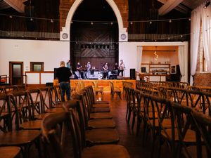 Empty banquet hall with wooden cross-back chairs arranged in rows facing a stage with spotlights and exposed brick walls.