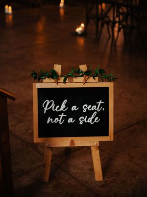 Wooden sign stands displaying 'Pick a seat, not a side' text at a wedding ceremony setup.
