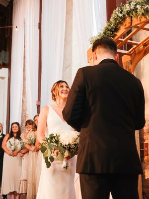 Wedding ceremony taking place in an elegant church interior with white drapery and floral decorations.
