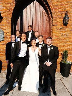 Wedding party in formal attire poses against rustic brick wall exterior of venue.