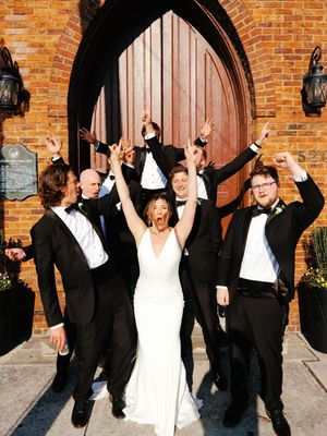 Wedding party celebrates with raised arms in front of rustic brick wall with arched wooden doors.
