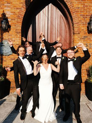 Wedding party jumping and celebrating outside a historic brick church entrance.