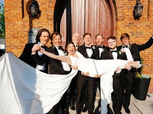 Wedding party celebrates with playful lift pose outside brick church entrance.
