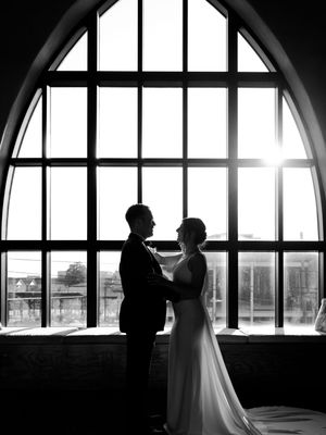 Silhouetted couple embraces in front of dramatic arched window with sunlight streaming through.