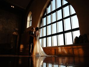 Dramatic backlit wedding photo against large arched window in historic venue.
