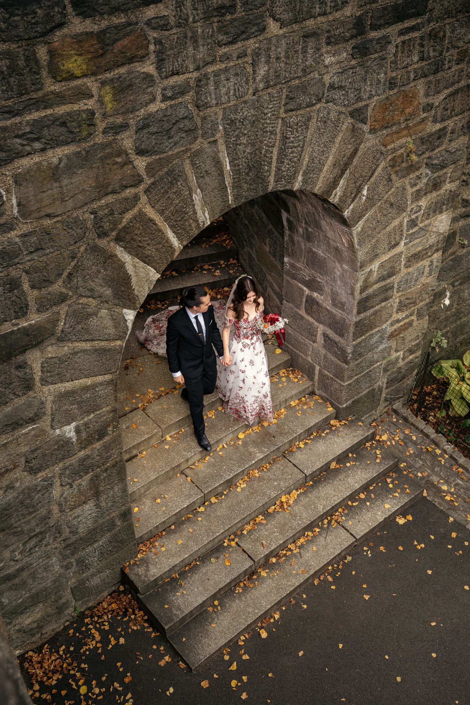 Bride and groom walk through a stone arch tunnel surrounded by autumn leaves at their wedding.