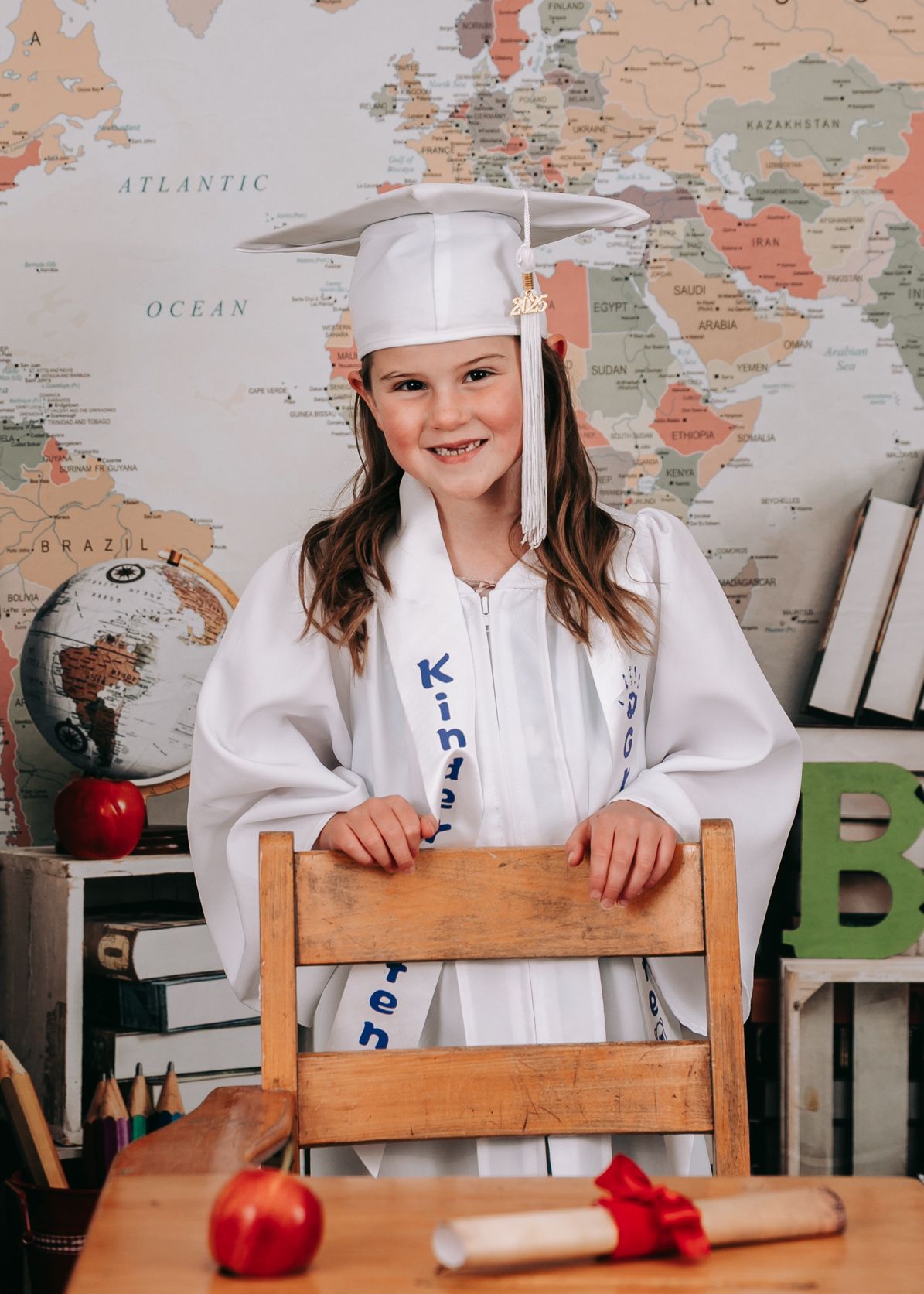 A smiling girl in a white kindergarten graduation cap and gown stands behind a wooden desk with a diploma.