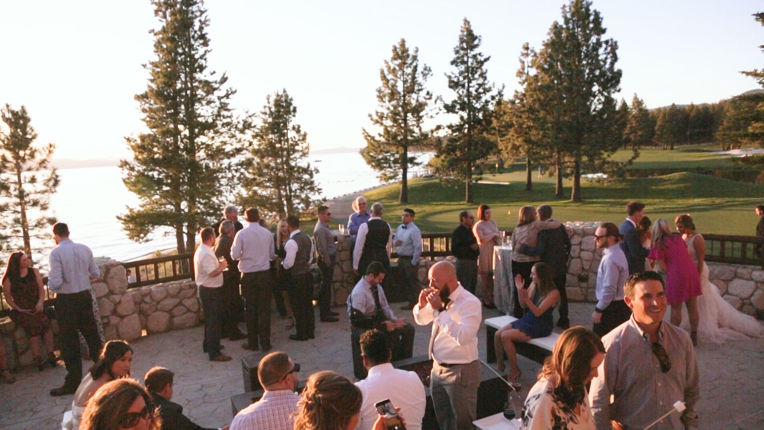 Guests mingle at an outdoor lakeside evening gathering surrounded by tall pine trees with scenic water views.