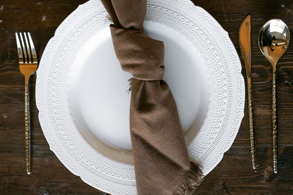 White embossed plate with brown linen napkin and gold cutlery on dark wood table.