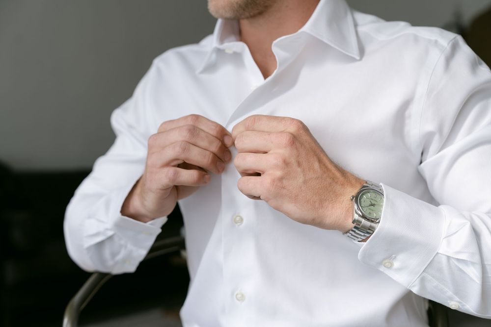 Man in white dress shirt buttoning collar, wearing silver watch, getting dressed for formal occasion.